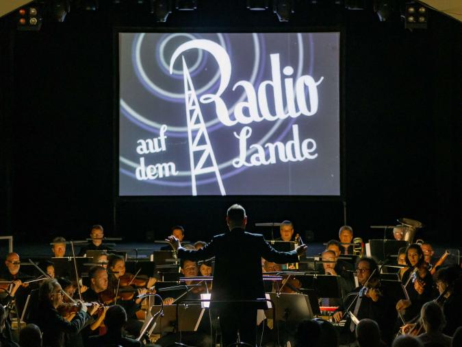 Orchester während eines Konzerts, im Hintergrund Leinwand mit Projektion „Radio auf dem Lande“