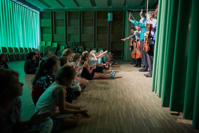 Im Kinderkonzert „Mitmach-Melodie der Großstadt“ sitzt eine Reihe Kinder vor den Mitgliedern der Kammerakademie Potsdam, die ihre Streichinstrumente in den Händen halten. Durch einen Vorhang scheint grünliches Licht.