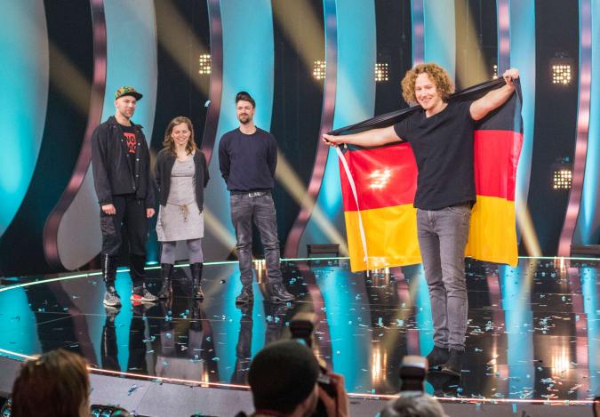 Der Gewinner des deutschen Vorentscheids zum Eurovision Song Contest 2018, Michael Schulte, steht auf der Bühne und spannt lachend eine Deutschlandfahne hinter seinem Rücken.