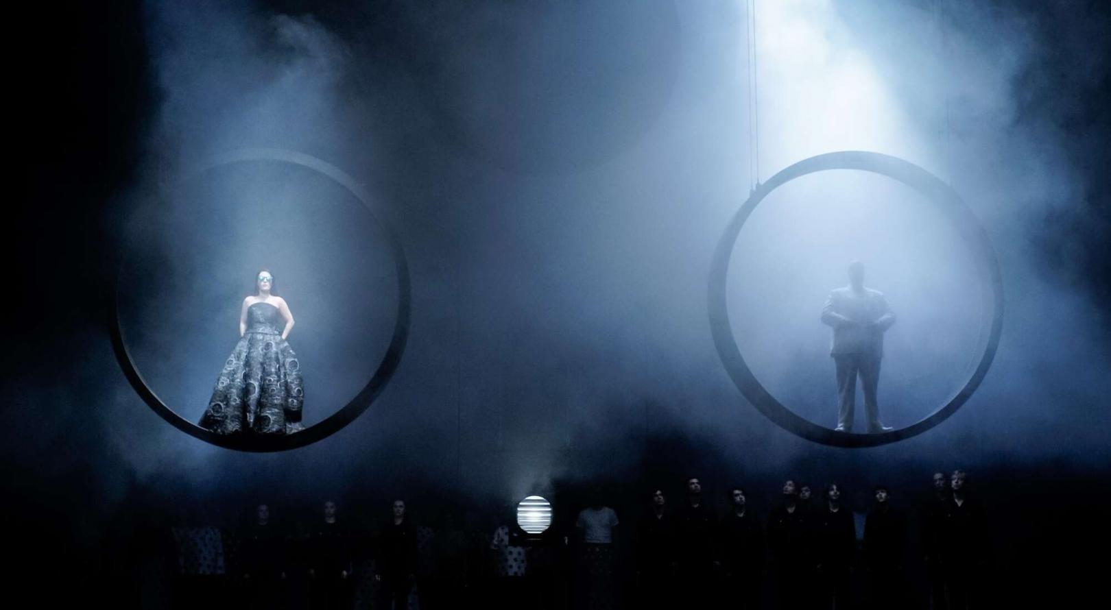 Szene auf einer Theaterbühne: männliche und weibliche Figur jeweils in einem Ring stehend schweben über der Bühne