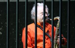 Älterer Herr in orangefarbenem Häftlingsoverall spielt Saxofon hinter Gittern