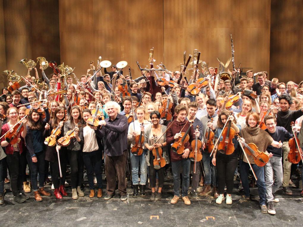 Eine große Gruppe junger Menschen mit Orchesterinstrumenten, davor Dirigent Sir Simon Rattle