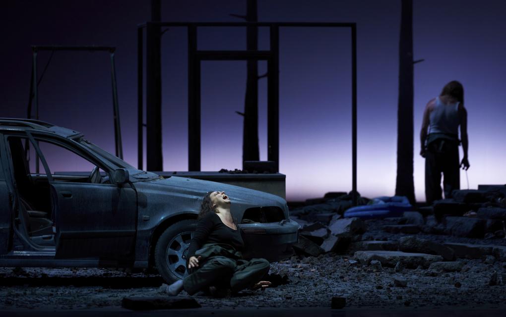 Szene auf einer Theaterbühne: Verzweifelte Frau sitzt neben Auto