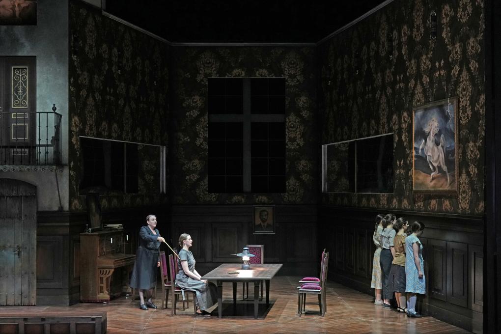 Szene auf einer Theaterbühne: Dame in schwarz zwingt sitzende Dame mit Stock, gerade zu sitzen. Vier junge Mädchen stehen mit dem Gesicht zur Wand.