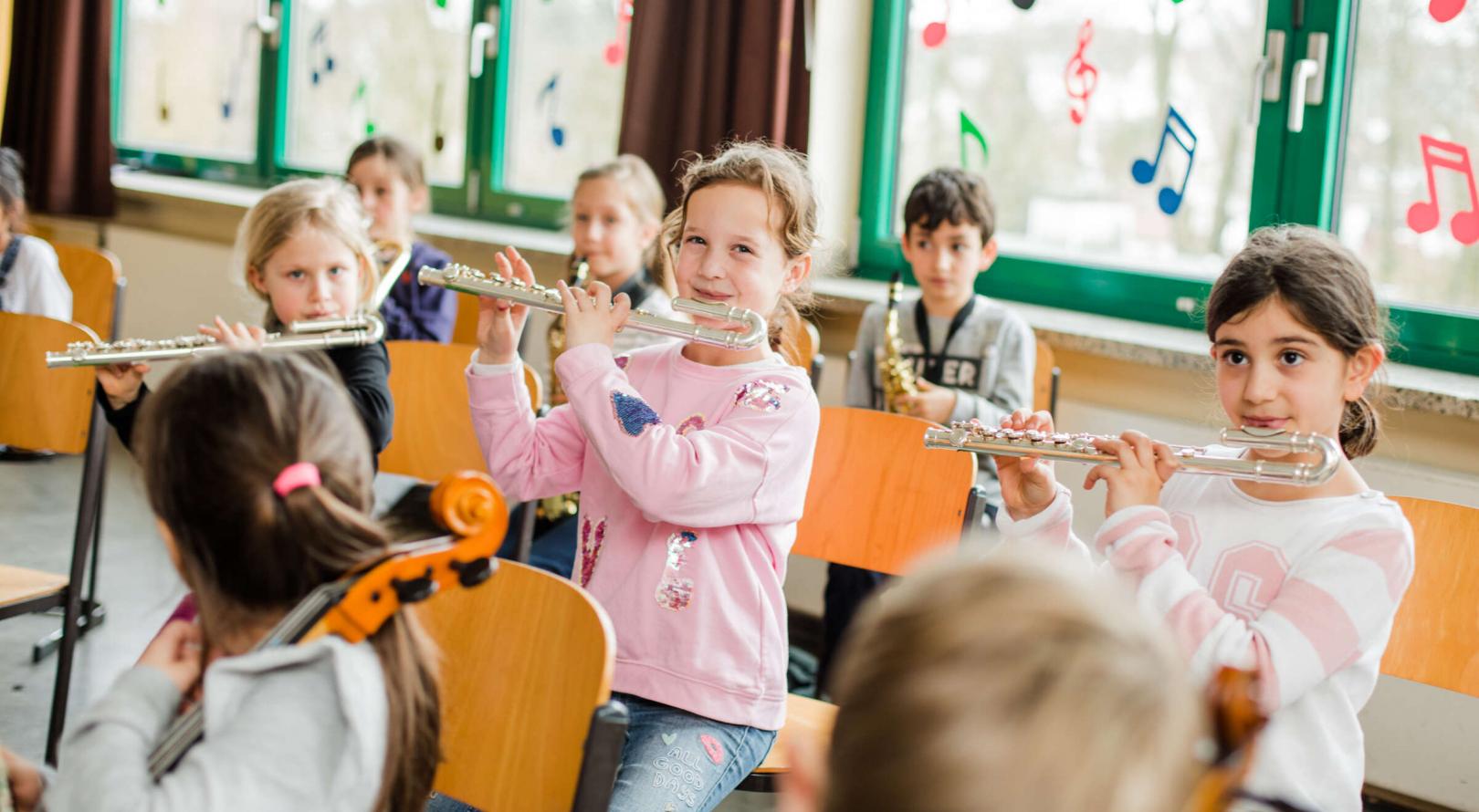 Kinder spielen gemeinsam im JeKits-Orchester auf ihren Instrumenten. Im Vordergrund des Fotos sind drei Mädchen mit ihren Kinder-Querflörten zu sehen.